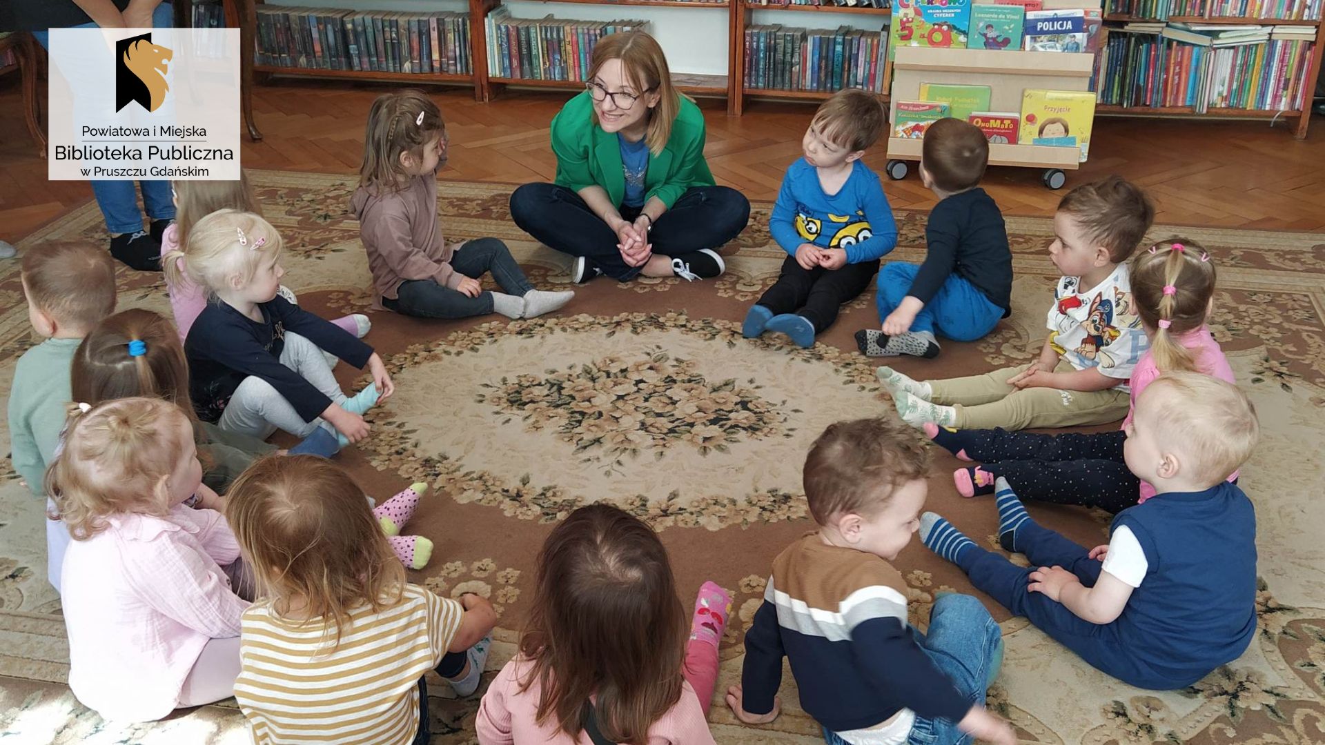 Dzieci wraz z panią bibliotekarką siedzą w okręgu na podłodze. W tle regały z książkami.