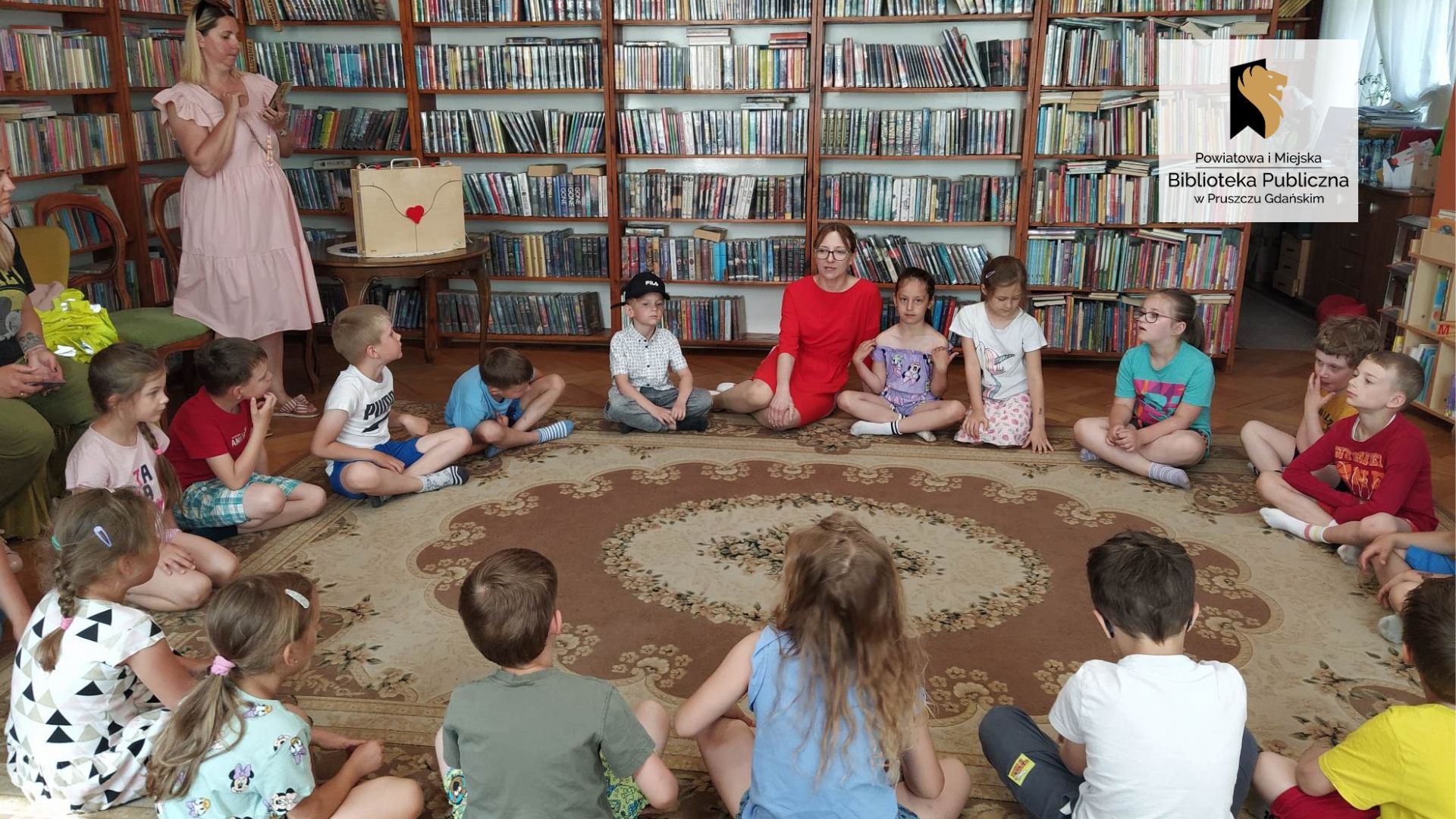 Dzieci wraz z panią bibliotekarką siedzą na podłodze w okręgu. Po lewej pani przedszkolanka robi zdjęcie. W tle regały z książkami.