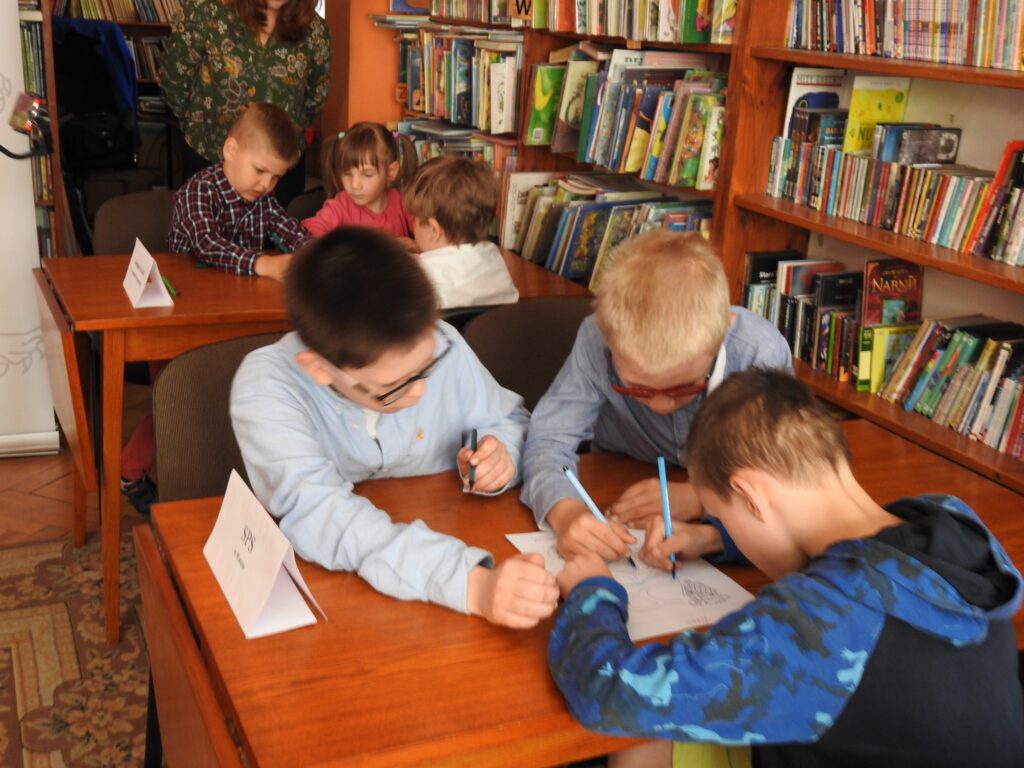 W sali bibliotecznej przy 2 stolikach siedzą dzieci. Przy każdym po troje. Dzieci wykonują zadanie konkursowe na kartkach.