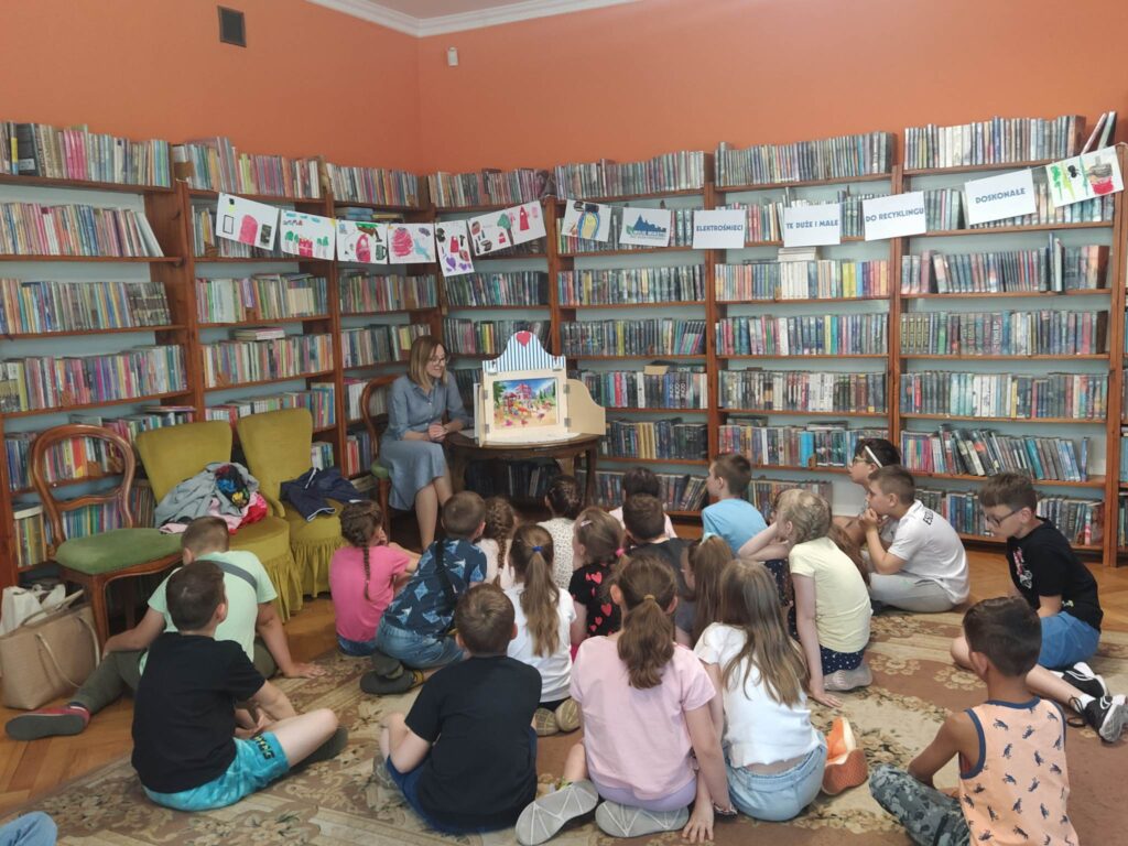 Dzieci siedzą tyłem i patrzą na teatrzyk kamishibai. Teatrzyk stoi na stole. Na kartce w teatrzyku znajduje się kolorowa ilustracja przedstawiająca dzieci na placu zabaw. Po lewej na krześle siedzi pani bibliotekarka i czyta. W tle regały z książkami.