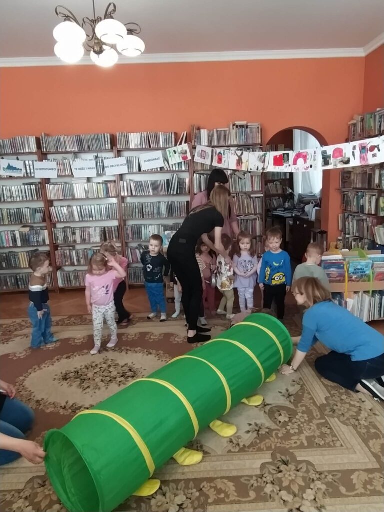 Na podłodze leży zielony tunel. Dzieci ustawiają się w kolejce, aby przez niego przejść. Między dziećmi stoją panie opiekunki. Pani bibliotekarka jest przy wejściu do tunelu. W tle regały z książkami. 