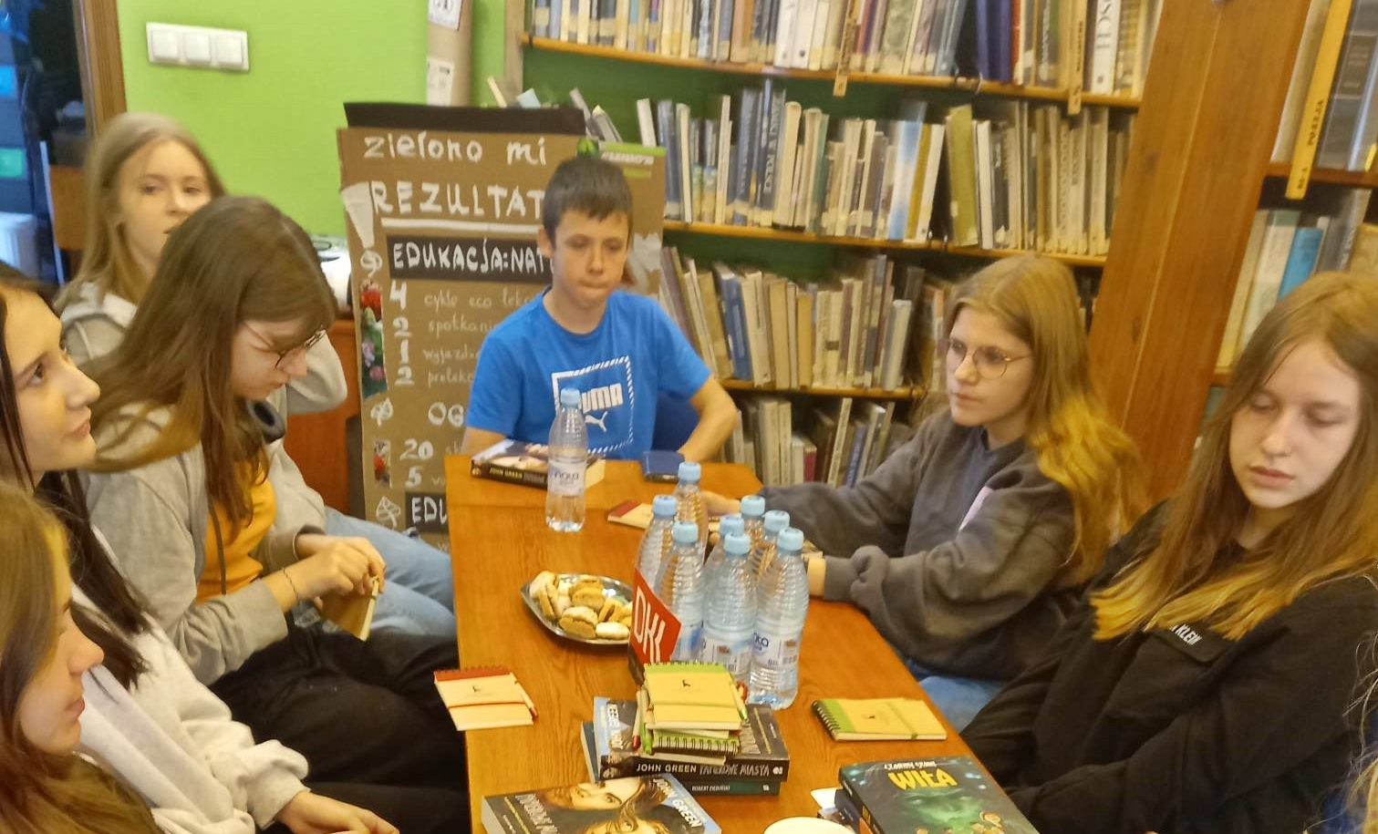 6 dziewczyn i 1 chłopiec z Dyskusyjnego klubu Książki siedzi przy stole. Na stole leżą Książki oraz stoją wody i ciastka.