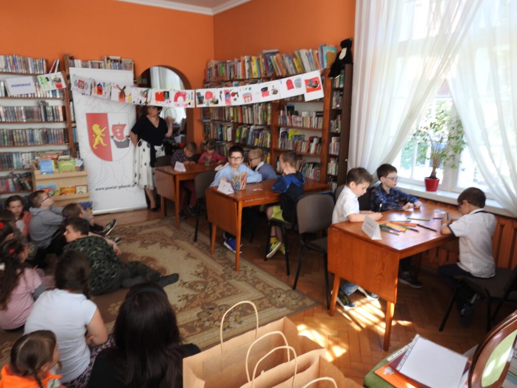 Przy 3 stołach siedzą w ,,trójkach" dzieci biorące udział w konkursie. Obok stoi inicjatorka oraz prowadząca konkurs Anna Krawycińska.