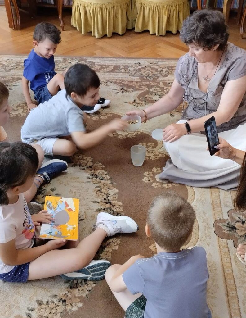 Dzieci siedzą na dywanie przed panią Anią, która trzyma w ręku miseczkę z kamykami.Chłopiec wyciąga rękę po kamyk. Dziewczynka ma rozłożona między nogami książkę. 