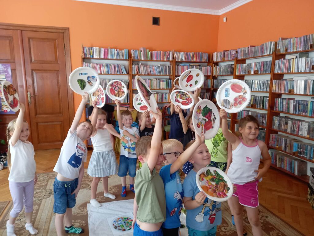 Dzieci stojąc trzymają w rękach białe talerze, na których naklejone są obrazki z owocami i warzywami.