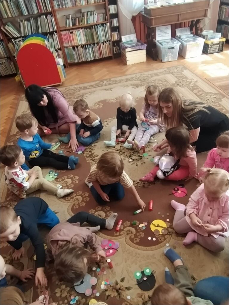 Dzieci wraz z opiekunkami siedzą na podłodze. Dzieci wykonują prace plastyczne. Przed nimi leżą materiały papiernicze. W tle regały z książkami.

