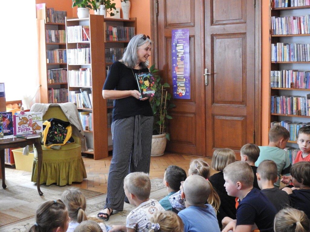 Roksana Jędrzejewska-Wróbel stoi przed dziećmi, które siedzą na podłodze. W ręce trzyma książkę i mówi. W tle regały z książkami oraz stolik, na którym stoją książki autorki.