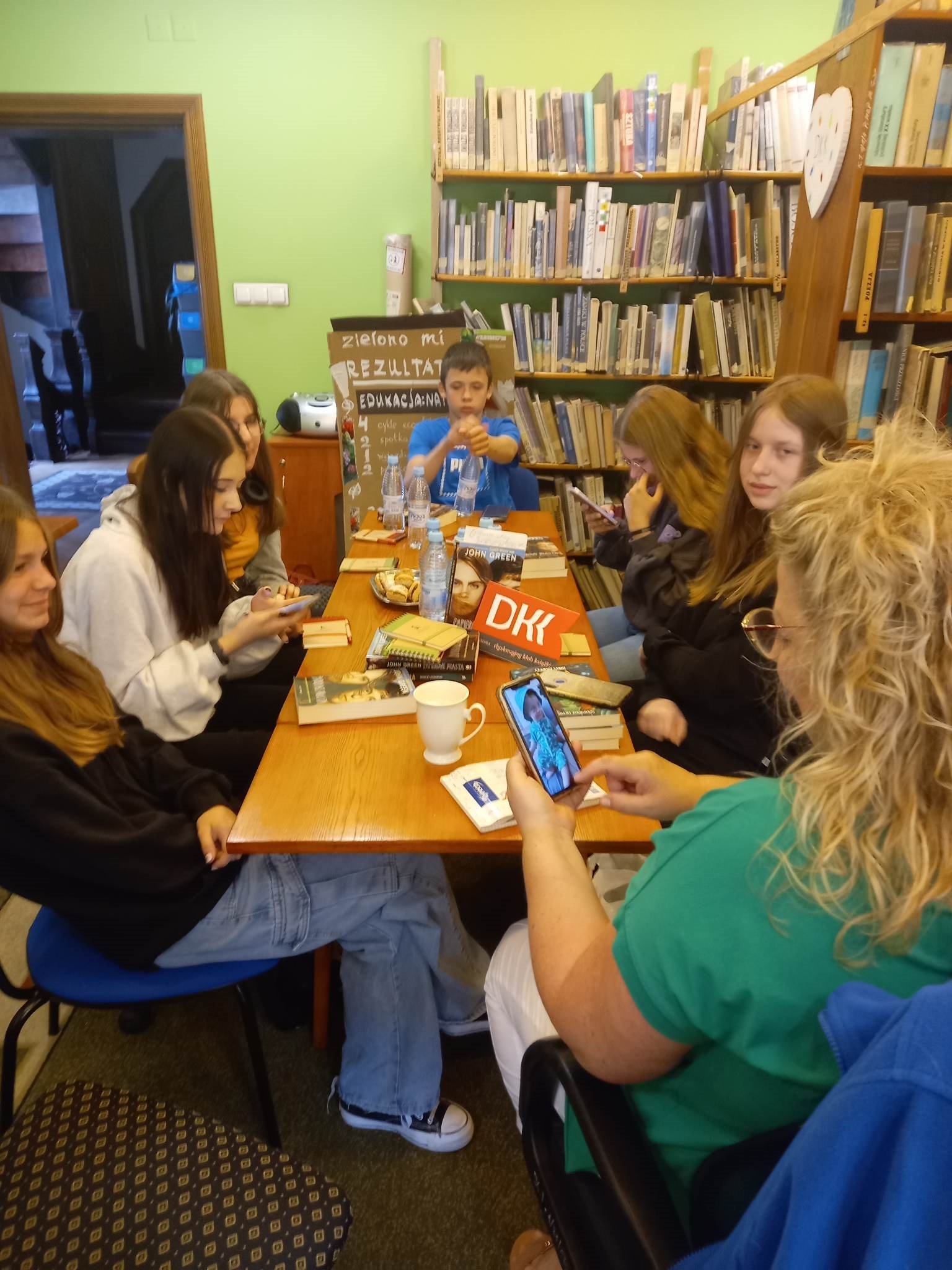 6 dziewczyn i 1 chłopiec z Dyskusyjnego klubu Książki siedzi przy stole. Z przodu stołu siedzi moderatorka klubu. Na stole leżą Książki oraz stoją wody i ciastka.