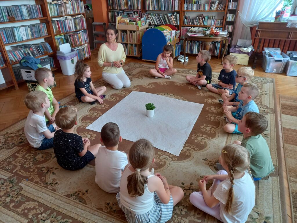 Na dywanie w okręgu siedzą dzieci i Pani Edyta Szordykowska. Pośrodku leży biały obrus, na którym stoi doniczka z natką pietruszki.