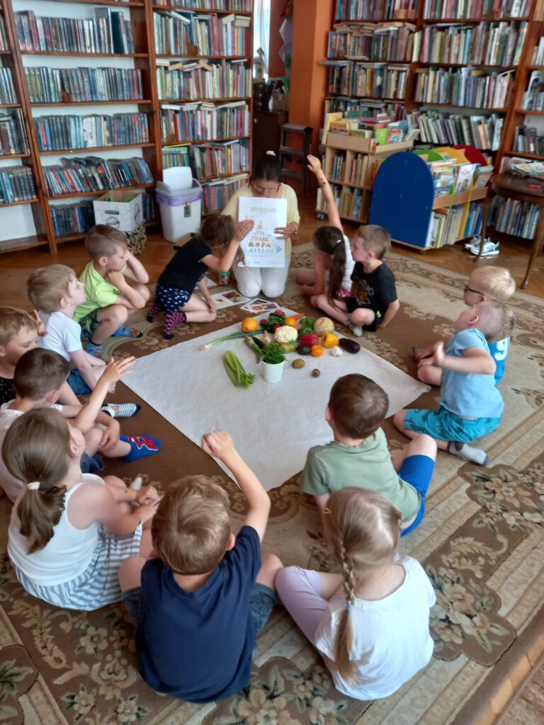 Dzieci siedzą na dywanie. Przed nimi leży biały obrus, na którym znajdują się warzywa i owoce. Pani Edyta trzyma w ręku obrazek z piramidą żywienia.