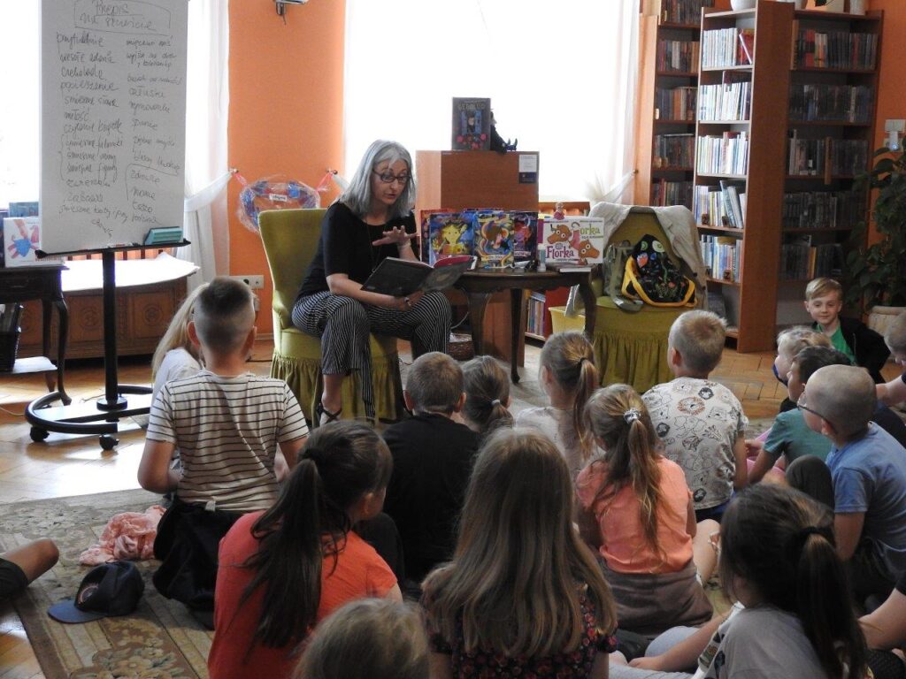 Roksana Jędrzejewska-Wróbel siedzi przy stoliku i czyta książkę. Na stoliku stoją książki autorki. Dzieci siedzą na podłodze i patrzą w jej stronę. W tle regały z książkami.