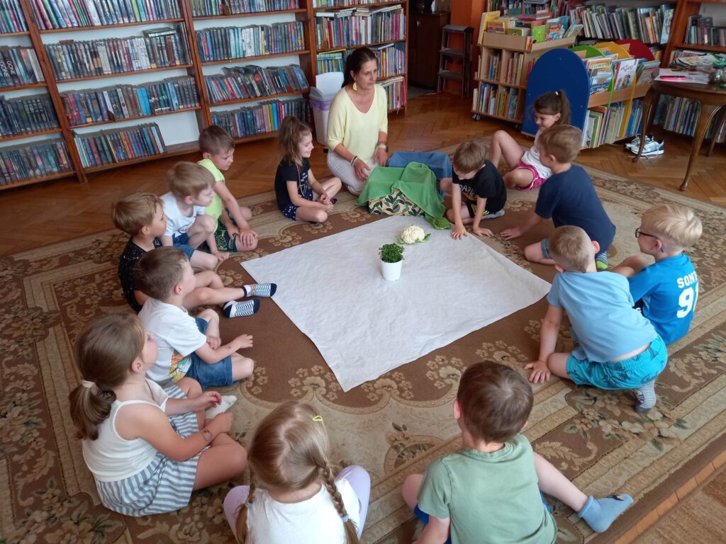 Na dywanie w okręgu siedzą dzieci i Pani Edyta Szordykowska. Pośrodku leży biały obrus, na którym stoi doniczka z natką pietruszki i leży kalafior. Obok Pani Edyty stoją dwa pudła.