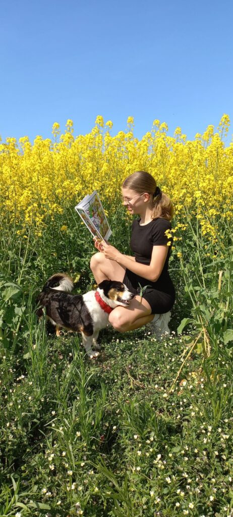 Na polu rzepaku siedzi dziewczynka i czyta książkę. na jej kolanach leży pies.