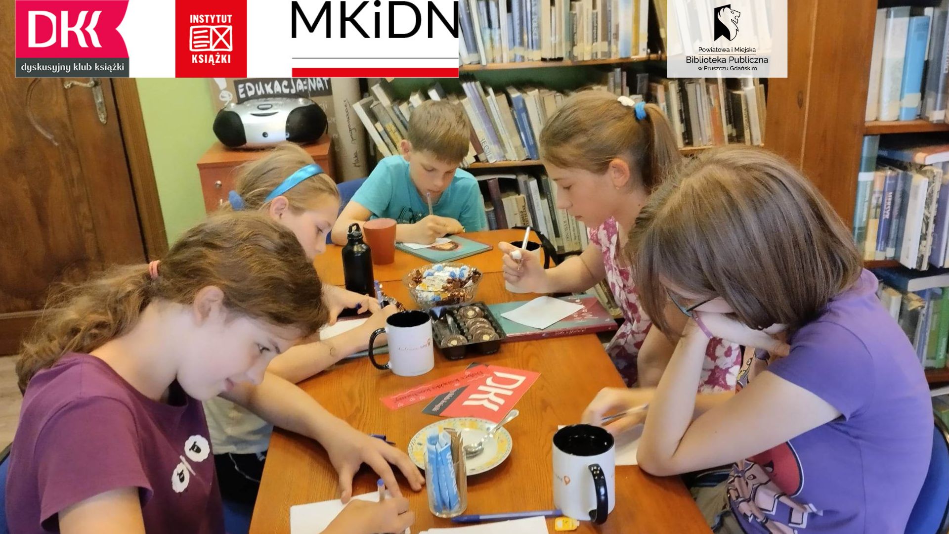 Przy stole siedzi młodzież. Przed sobą maja kartki. Po środku stołu leży logo DKK.