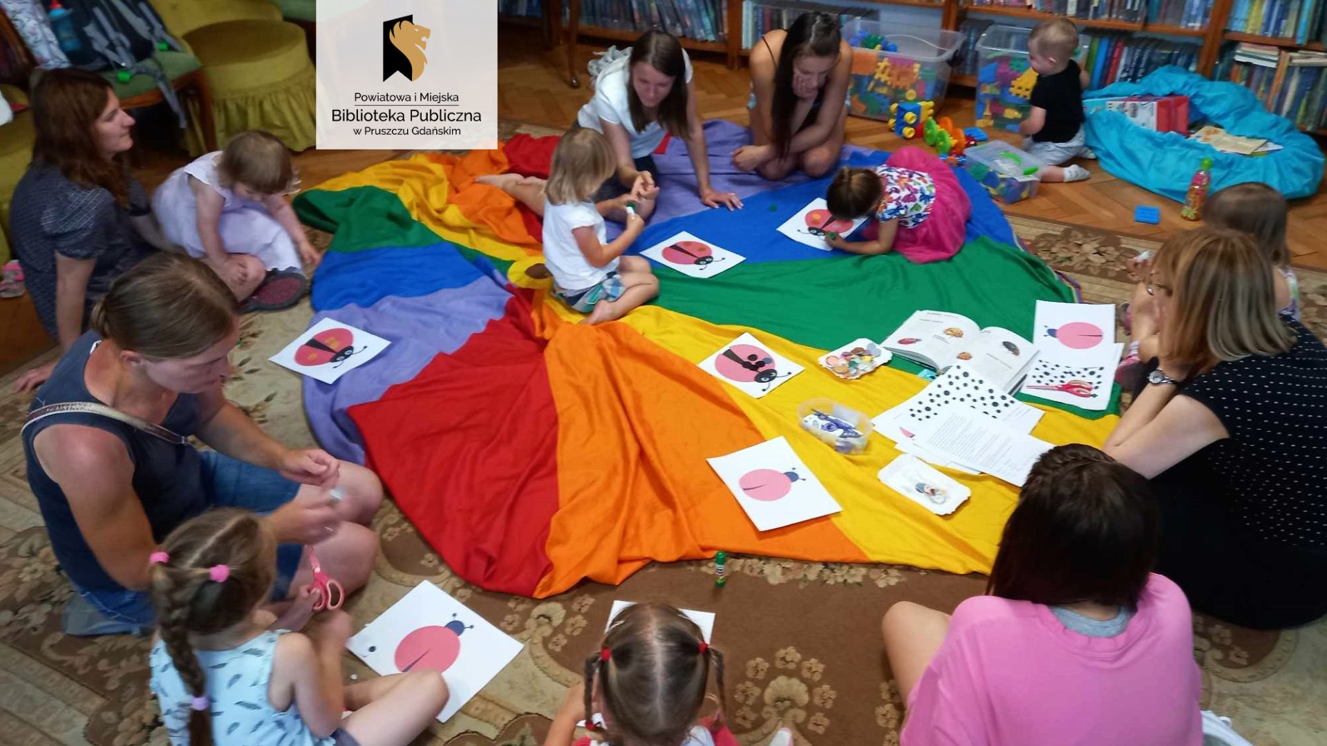 Wokół kolorowej chusty siedzą dzieci z mamami. Na chuście leżą obrazki z biedronkami. przy regałach z książkami stoją pudła z zabawkami.