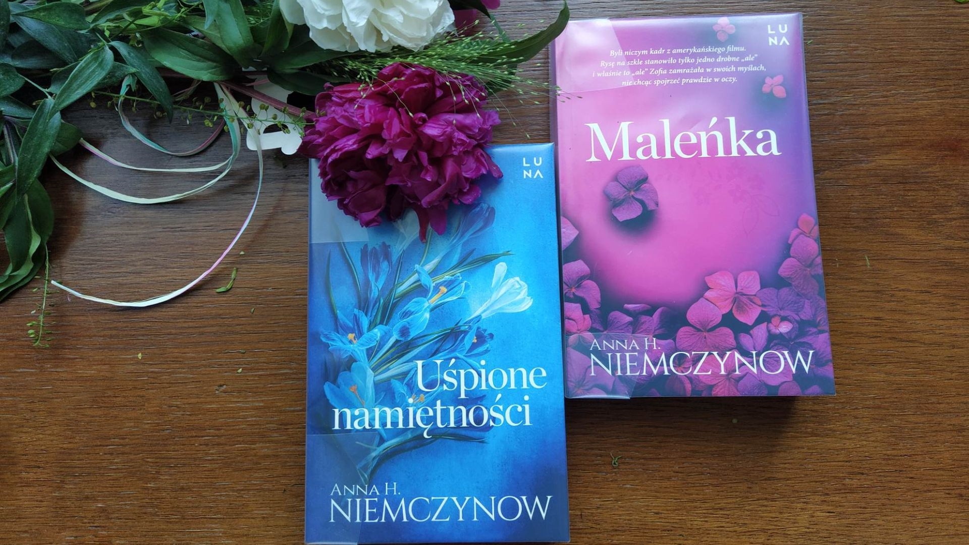 Dwie książki: „Maleńka” i „Uśpione namiętności” leża na drewnianym blacie. Nad nimi fioletowy kwiat.