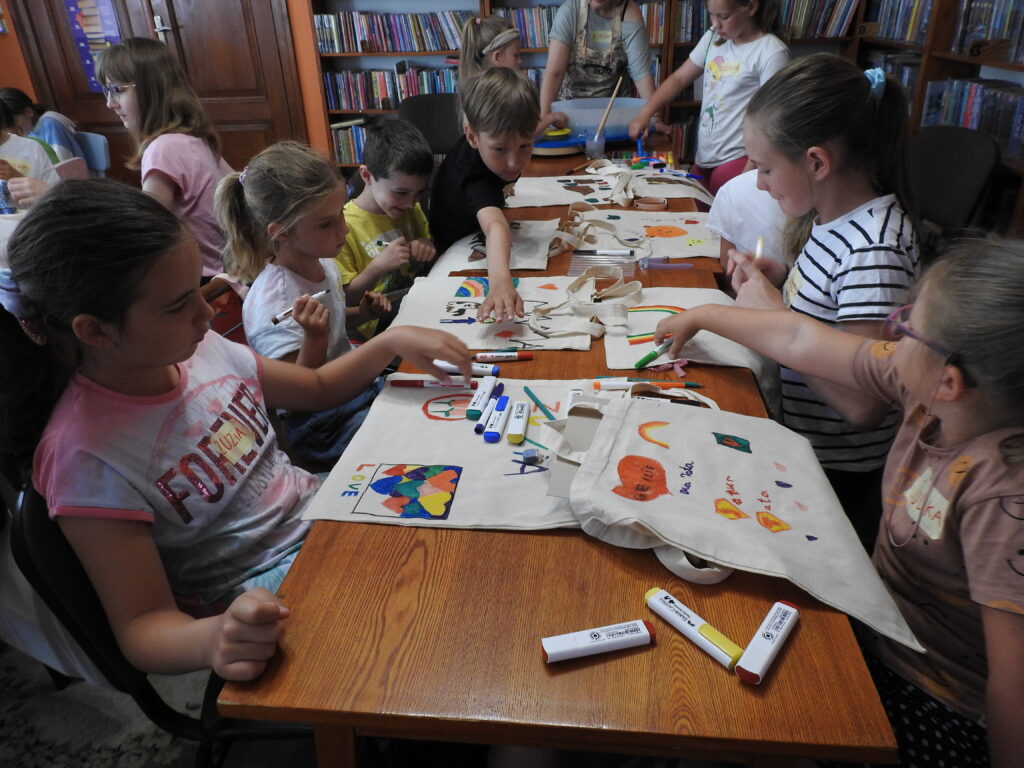 10 Dzieci siedzą przy stołach  i malują bawełniane torby. W tle regały z książkami.