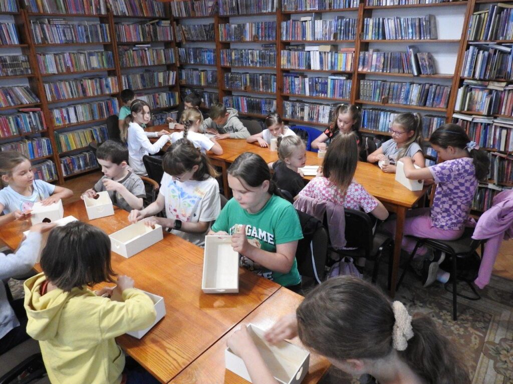 Dzieci siedzą przy stołach. Przed sobą mają drewniane skrzyneczki, które szlifują papierem ściernym. W tle regały z książkami.