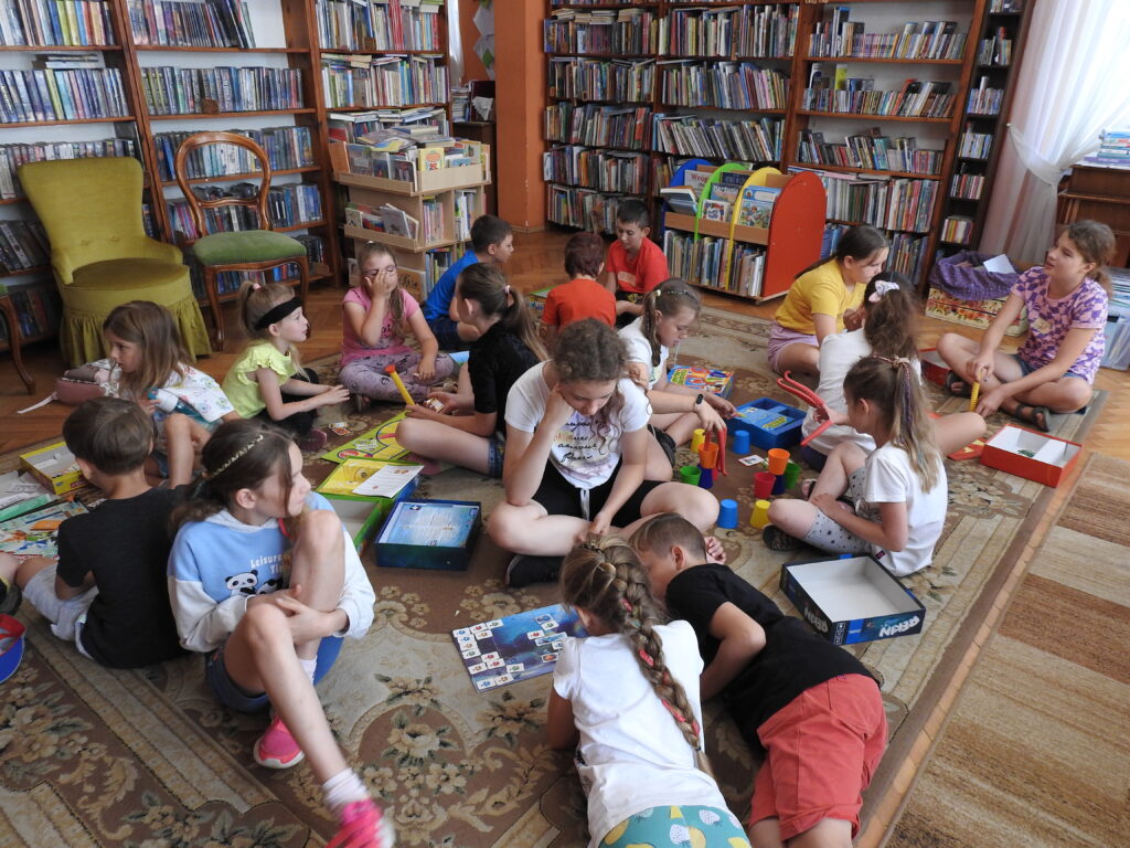 Dzieci siedzą na podłodze. Przed nimi leżą rozłożone różne gry planszowe W tle regały z książkami i krzesła.