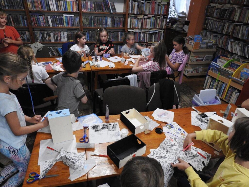 Dzieci siedzą przy stołach. Przed nimi leżą przybory papiernicze i drewniane skrzyneczki. Dzieci wykonują prace techniką decoupage. W tle regały z książkami.