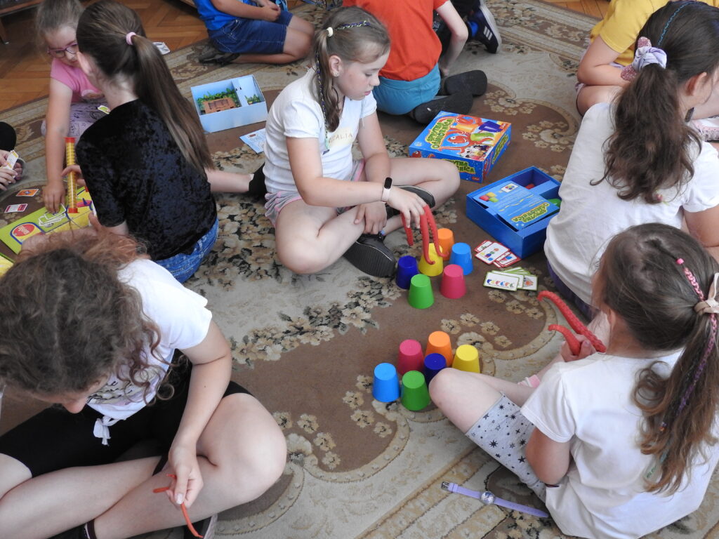 Dwie dziewczynki siedzą na podłodze i grają w grę Sprytna ośmiornica. Dziewczynki na palcach mają założone zabawkowe macki ośmiornicy. Przed nimi stoją kolorowe kubeczki. W tle inne dzieci grają w gry.