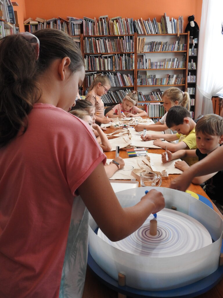 4 Dziewczynka używa maszyny kolorów,  trzyma kredkę w dłoni. W tle dzieci malują na bawełnianych torbach. Za nimi regały z książkami.