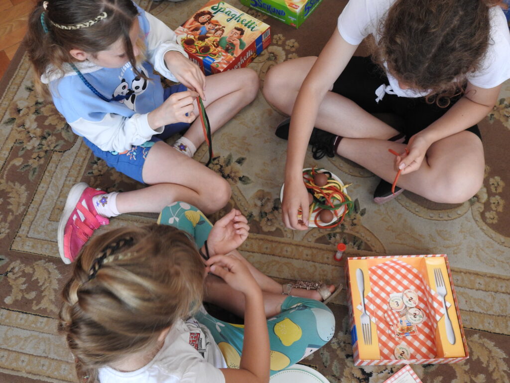 Trzy dziewczynki grają w grę Spaghetti. Przed sobą mają zabawkowy talerzyk i kolorowe sznurki imitujące makaron z klopsikami. Jedna z dziewczynek wyciąga kolorowy sznurek z talerza, a pozostałe zawiązują sznurki na rękach.
