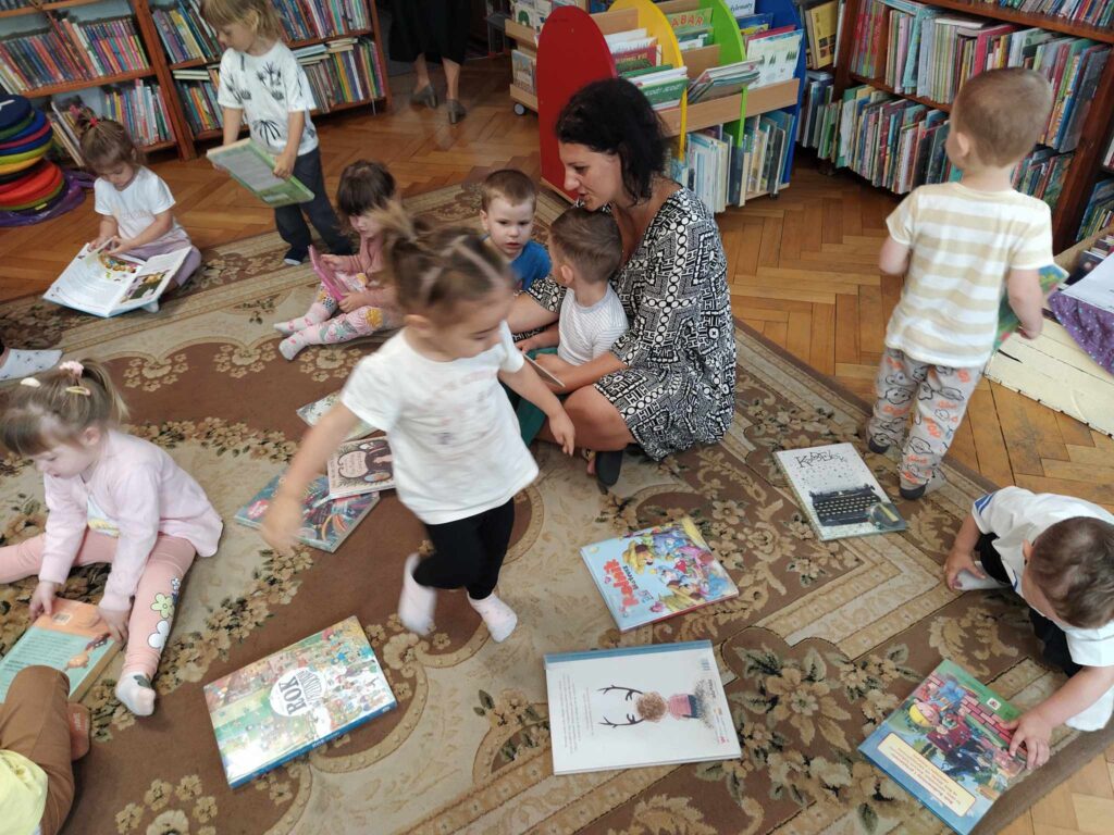 Dzieci siedzą na podłodze wraz z panią i oglądają książeczki. Niektóre z dzieci są w ruchu. W tle regały z książkami.