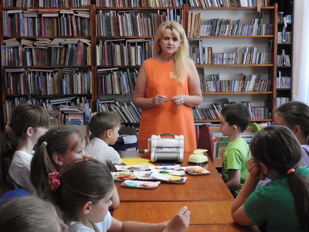 Maria Kierska – nauczycielka zaproszona na zajęcia, mówi do dzieci, które siedzą przy stole. Na stole materiały papiernicze, dziurkacz i wycinarka do scrapbookingu. W tle regały z książkami. 