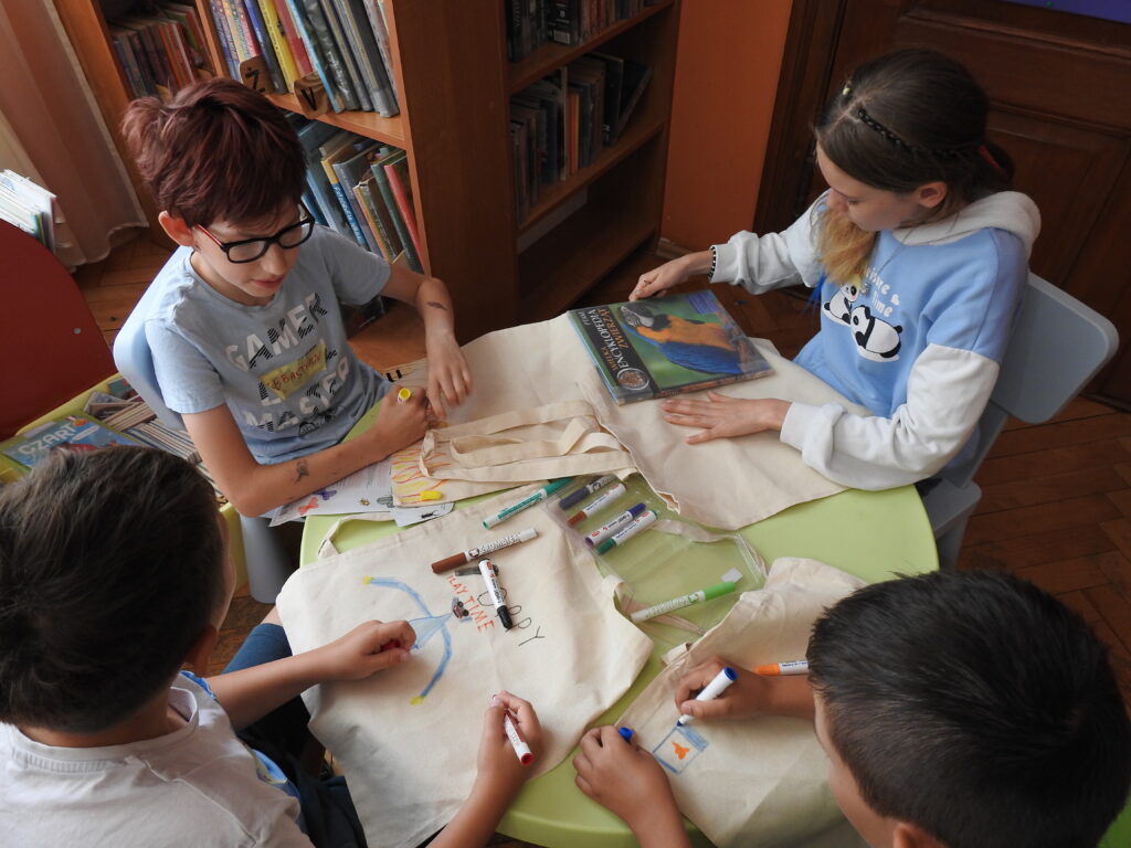 Czworo dzieci siedzi przy stoliku i maluje bawełniane torby. Dziewczynka trzyma książkę z papugą na okładce. W tle regały z książkami. 