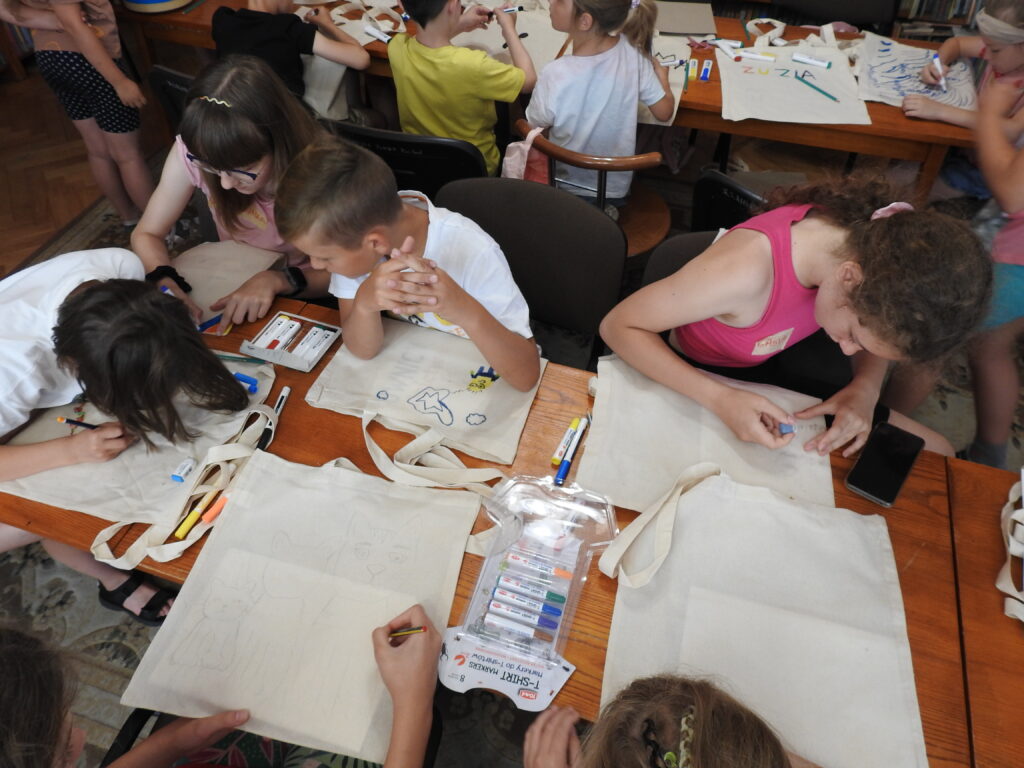 Dzieci siedzą przy stołach  i malują bawełniane torby. W tle regały z książkami.