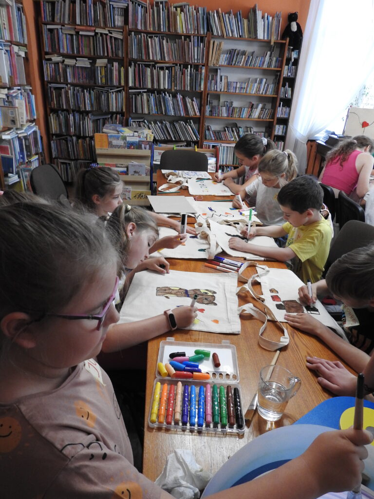 Dzieci siedzą przy stołach  i malują bawełniane torby. W tle regały z książkami.