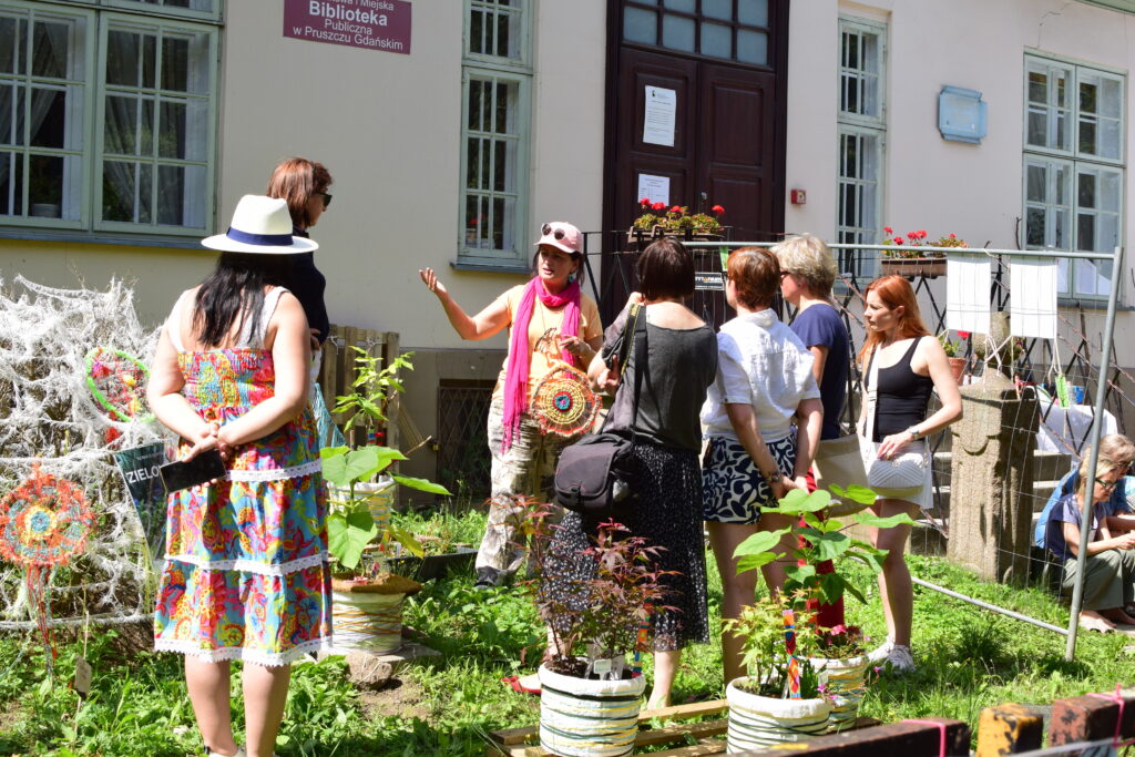 Grupa osób stoi przed budynkiem biblioteki, gdzie znajduje się mobilny ogród i słucha prezeski Fundacji KIWI, która opowiada o znajdujących się w nim roślinach.