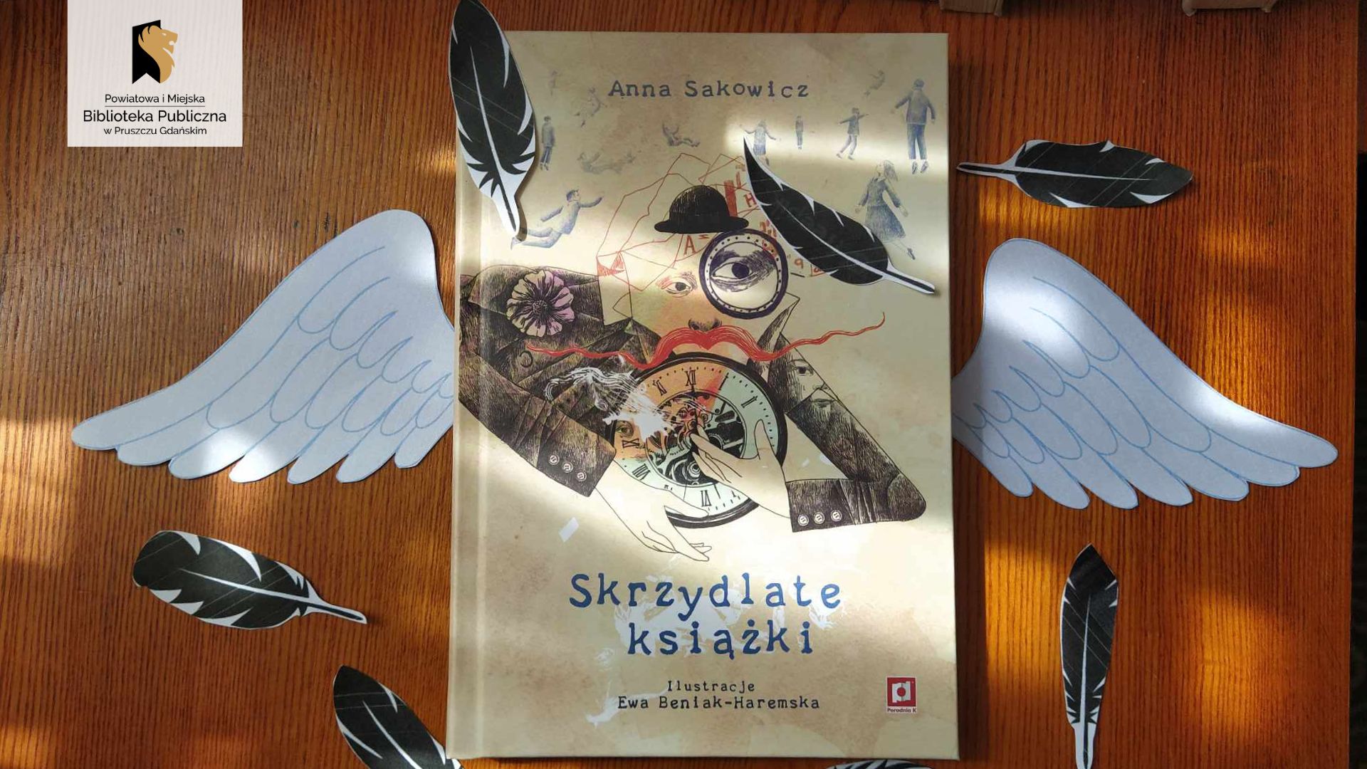 Na brązowym tle leży książka Anny Sakowicz pt. „Skrzydlate książki". Po obu stronach książki leżą papierowe skrzydła i czarne pióra.