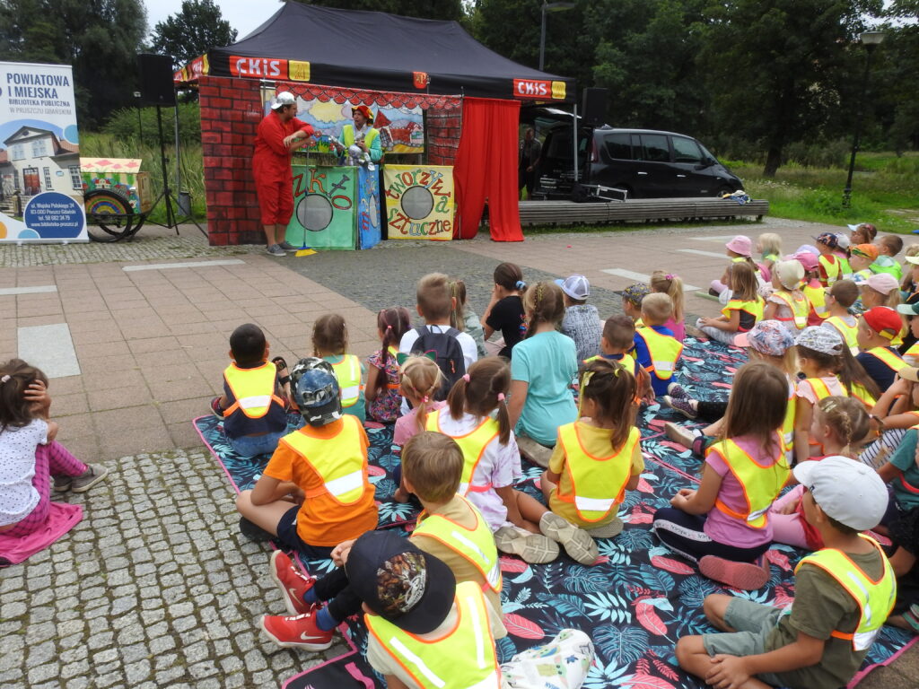 Duża grupa dzieci siedzi na świeżym powietrzu z głowami zwróconymi w stronę sceny teatralnej. Na scenieaktorzy ubrani w kolorowe stroje.