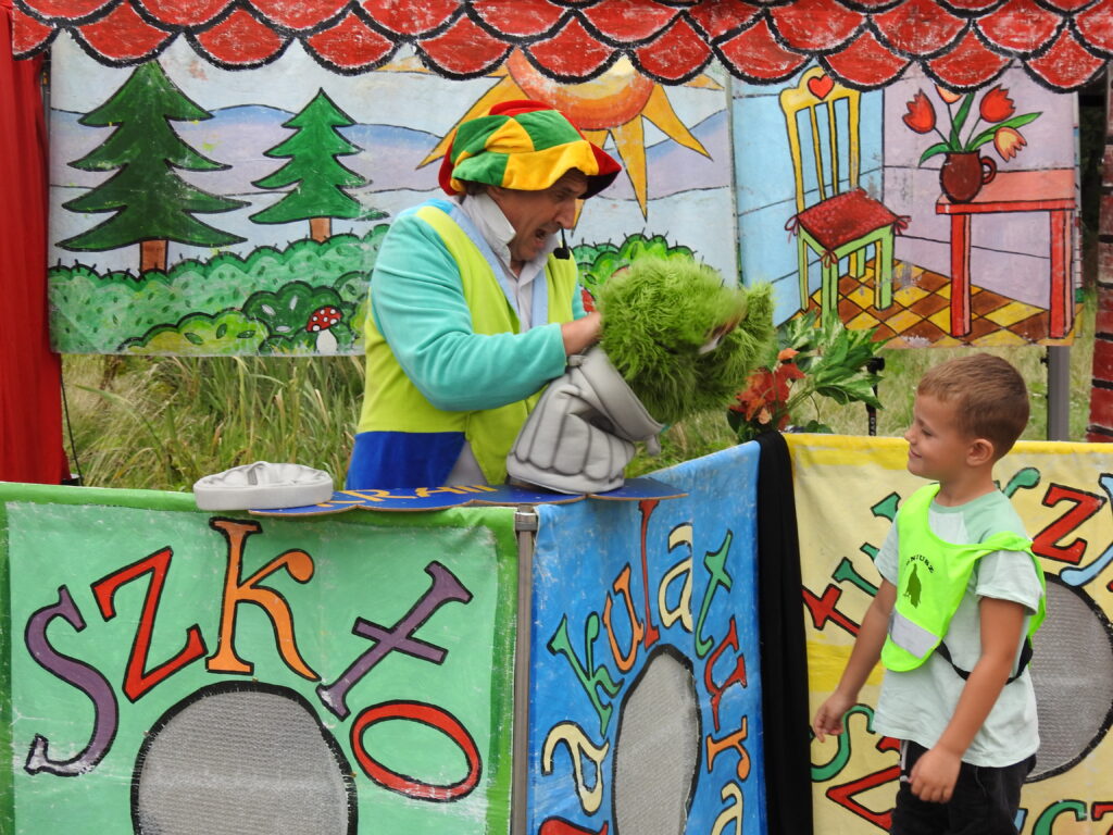 Aktor  w zielonym stroju i kolorowym kapeluszu stoi na scenie. Przed nim stoi dziecko, patrzące na zieloną zabawkę, którą trzyma aktor w rękach.