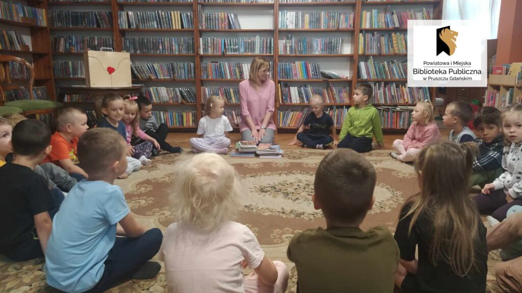 Dzieci wraz z panią bibliotekarką siedzą w okręgu na podłodze. Po lewej stronie na stoliku stoi teatrzyk kamishibai. W tle regały z książkami.