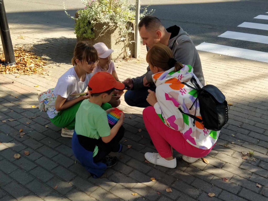 Uczestnicy gry miejskiej. 3 dzieci i 2 dorosłych kuca na chodniku