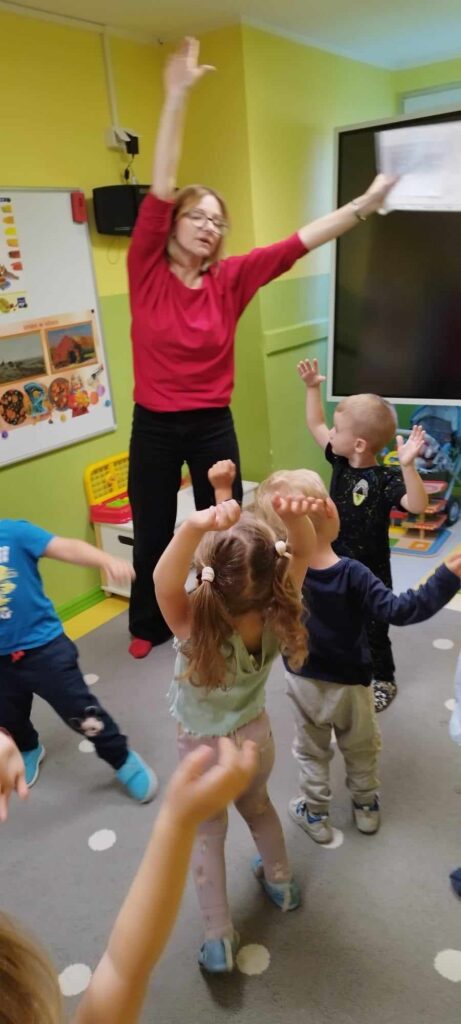 W sali przedszkolnej grupka małych dzieci i bibliotekarka. Dzieci powtarzają ruchy bibliotekarki, która wymachuje rękoma. 