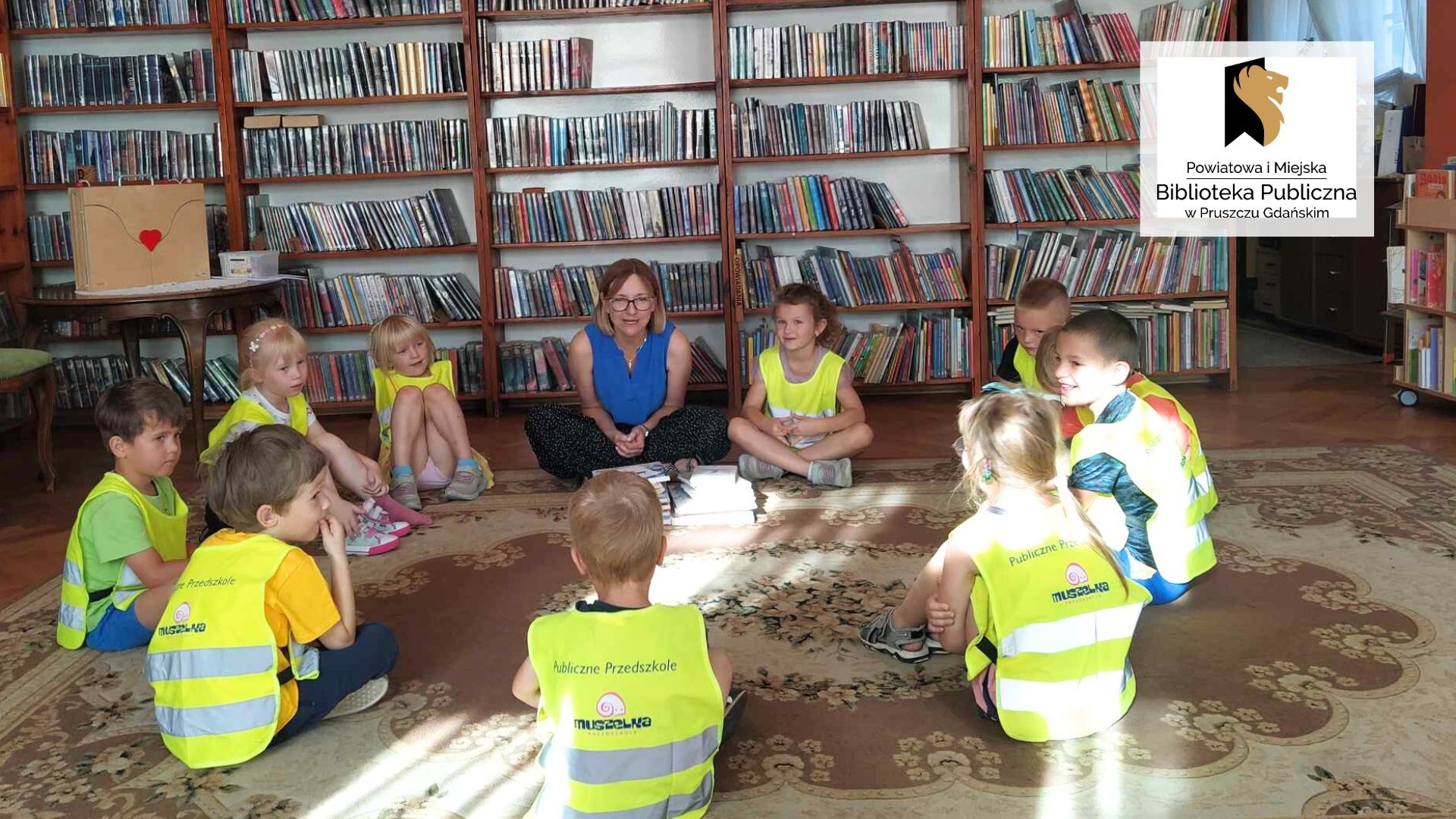Dzieci wraz z panią bibliotekarką siedzą w okręgu na podłodze. Po lewej na stoliku stoi teatrzyk kamishibai. W tle regały z książkami.