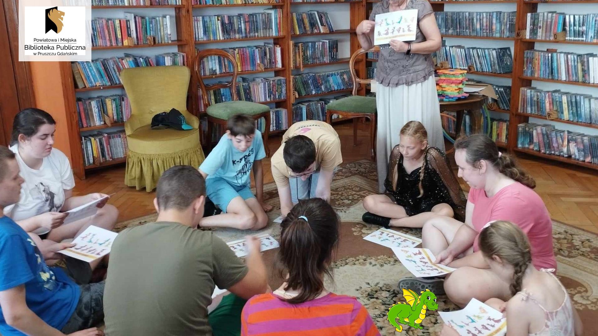 9 nastoletnich dzieci siedzi na dywanie w wypożyczalni dla dzieci. Przed nimi leżą kartki, na których wydrukowane są głowy smoka. Obok nich stoi Anna Krawycińska, prowadząca zajęcia, która trzyma w rękach taką samą kartę i cos objaśnia.