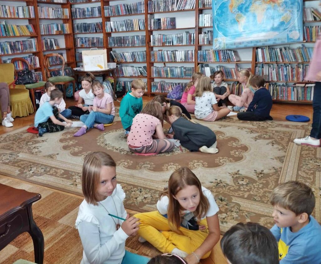 Dzieci uczestniczące w lekcji bibliotecznej siedzą na dywanie w grupkach i coś piszą.