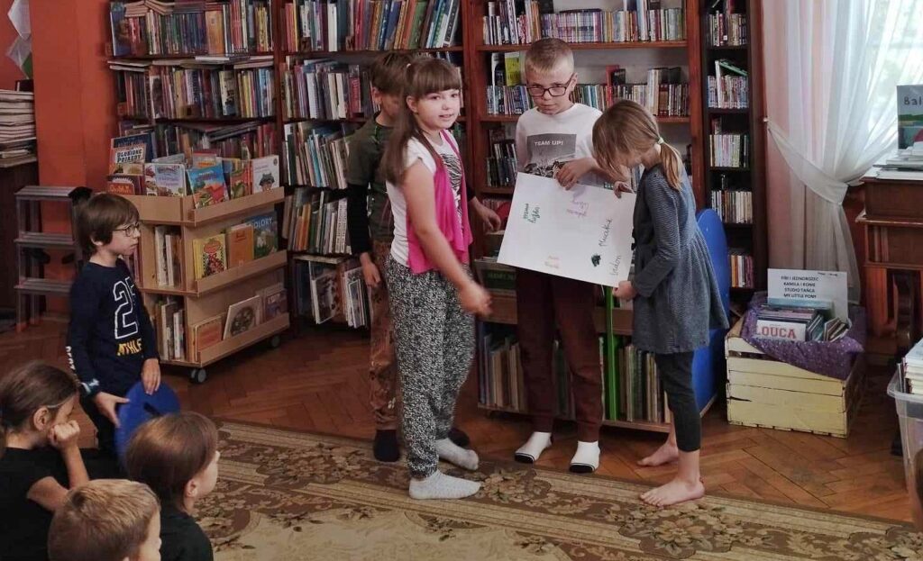 Trójka dzieci biorących udział w lekcji bibliotecznej stoi i prezentuje swoją pracę. Dzieci trzymają w rekach kartkę, na której jest coś napisane