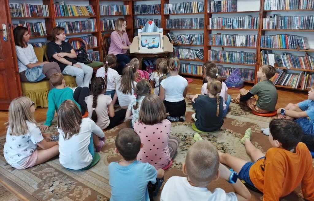 Duża grupa dzieci siedzi na dywanie w bibliotece. Dzieci patrzą pna bibliotekarkę, która czyta teatrzyk kamishibai pt. Szkoła Czi -Tam.