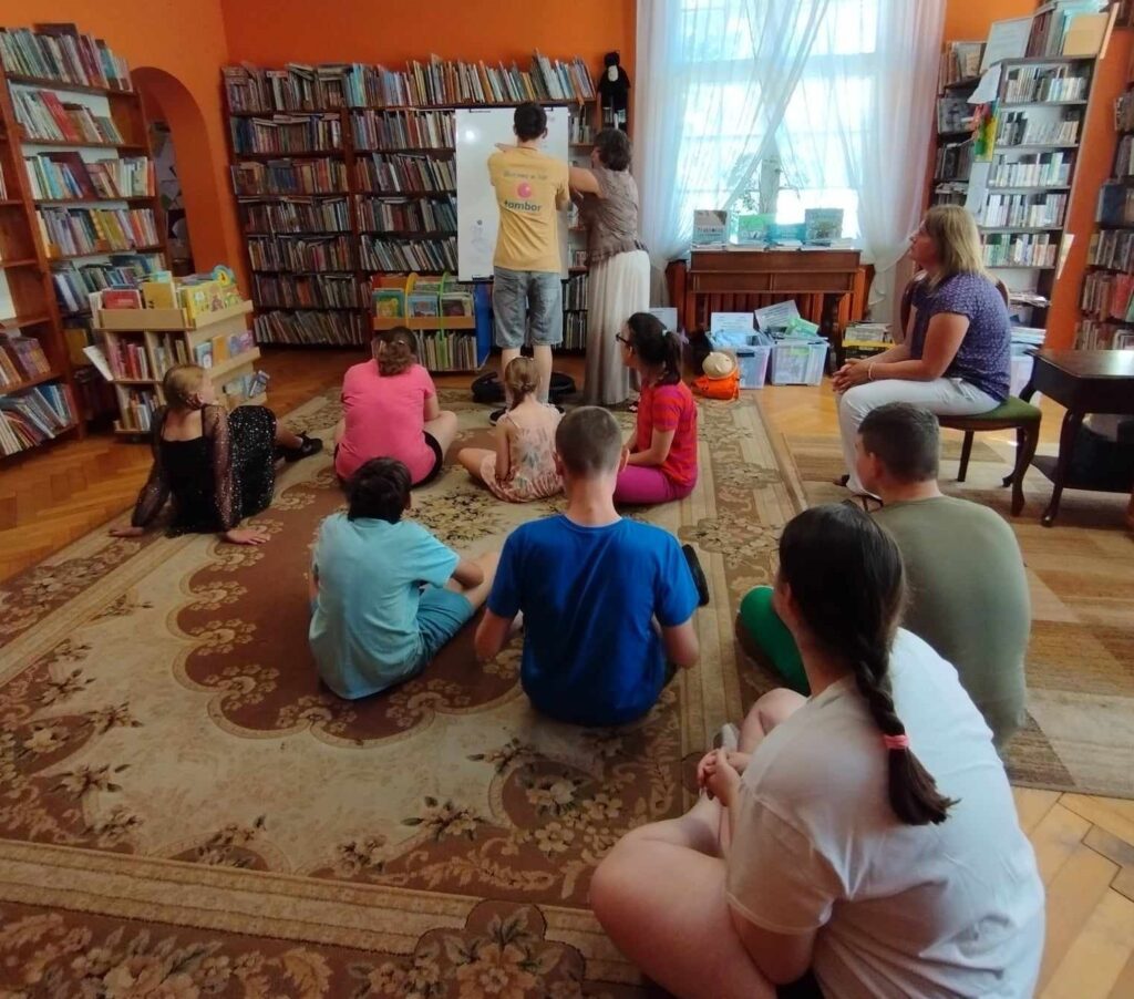 9 dzieci siedzi na dywanie w sali bibliotecznej. Dzieci patrzą w kierunku bibliotekarki i chłopca stojącego przy białej tablicy. 