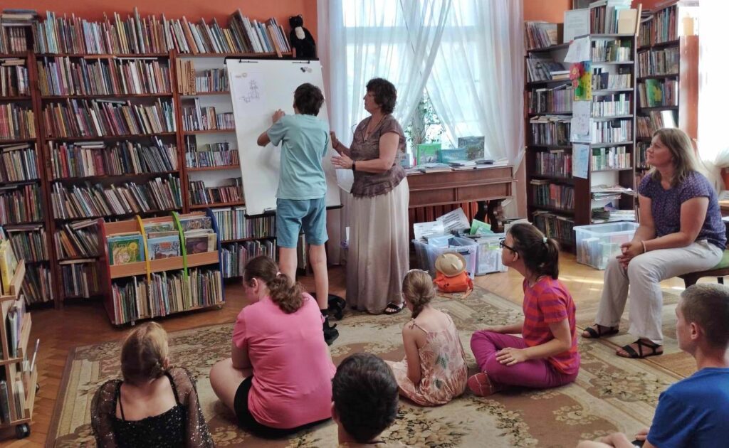 9 dzieci siedzi na dywanie w sali bibliotecznej. Dzieci patrzą w kierunku bibliotekarki i chłopca stojącego przy białej tablicy. 