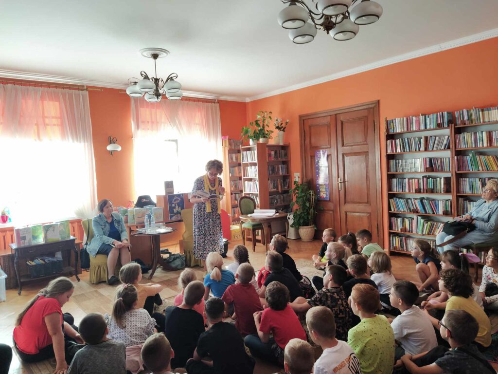 Dzieci siedzą na podłodze. Przed nimi stoi Malwina Kożurno i czyta książkę. Za nią ,w fotelu przy stoliku, siedzi Renata Piątkowska. Za nią książki i okna. Po prawej regały z książkami.