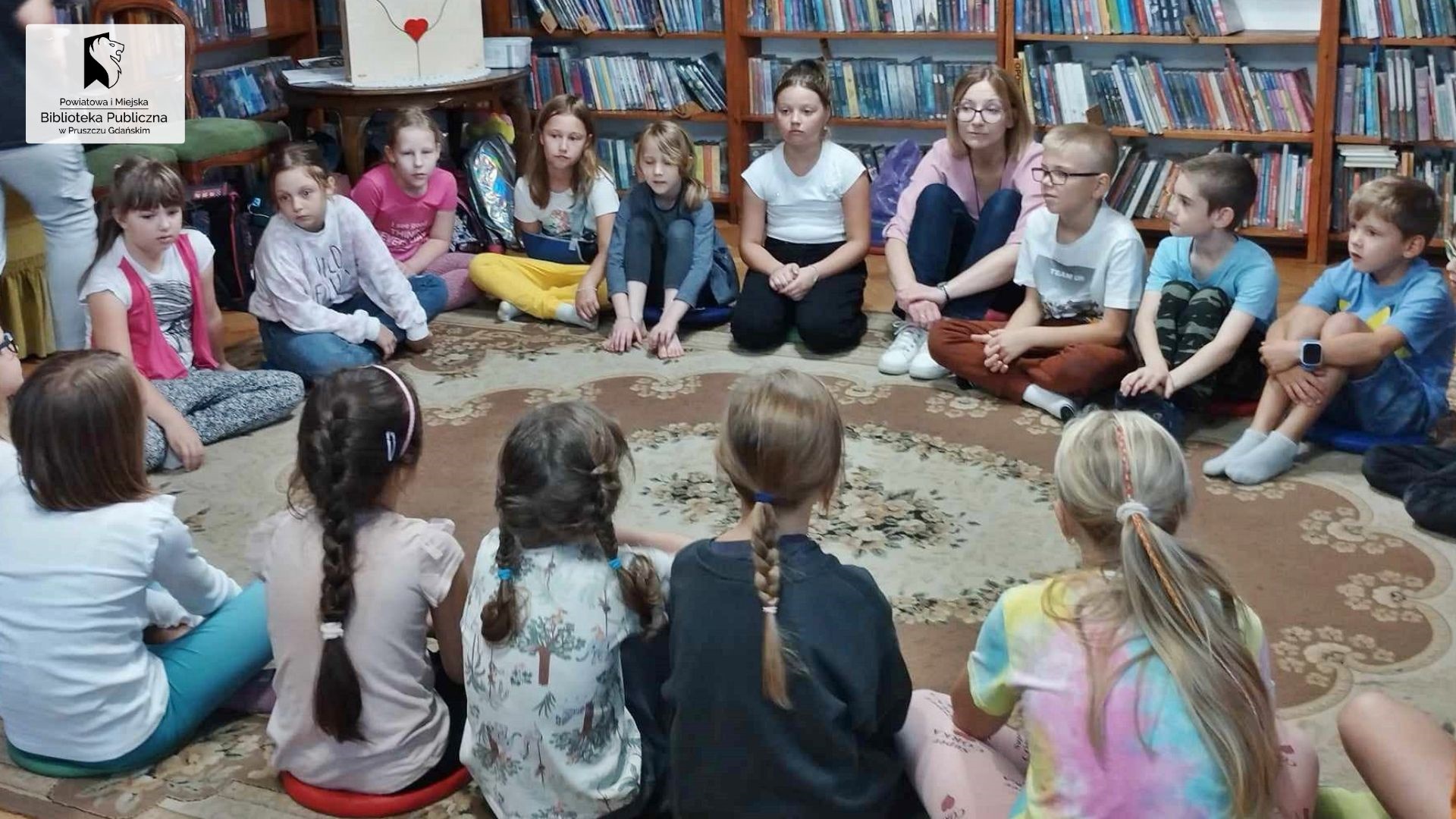 Duża drupa dzieci siedzi na dywanie w bibliotece. Między nimi siedzi bibliotekarka, która coś mówi.