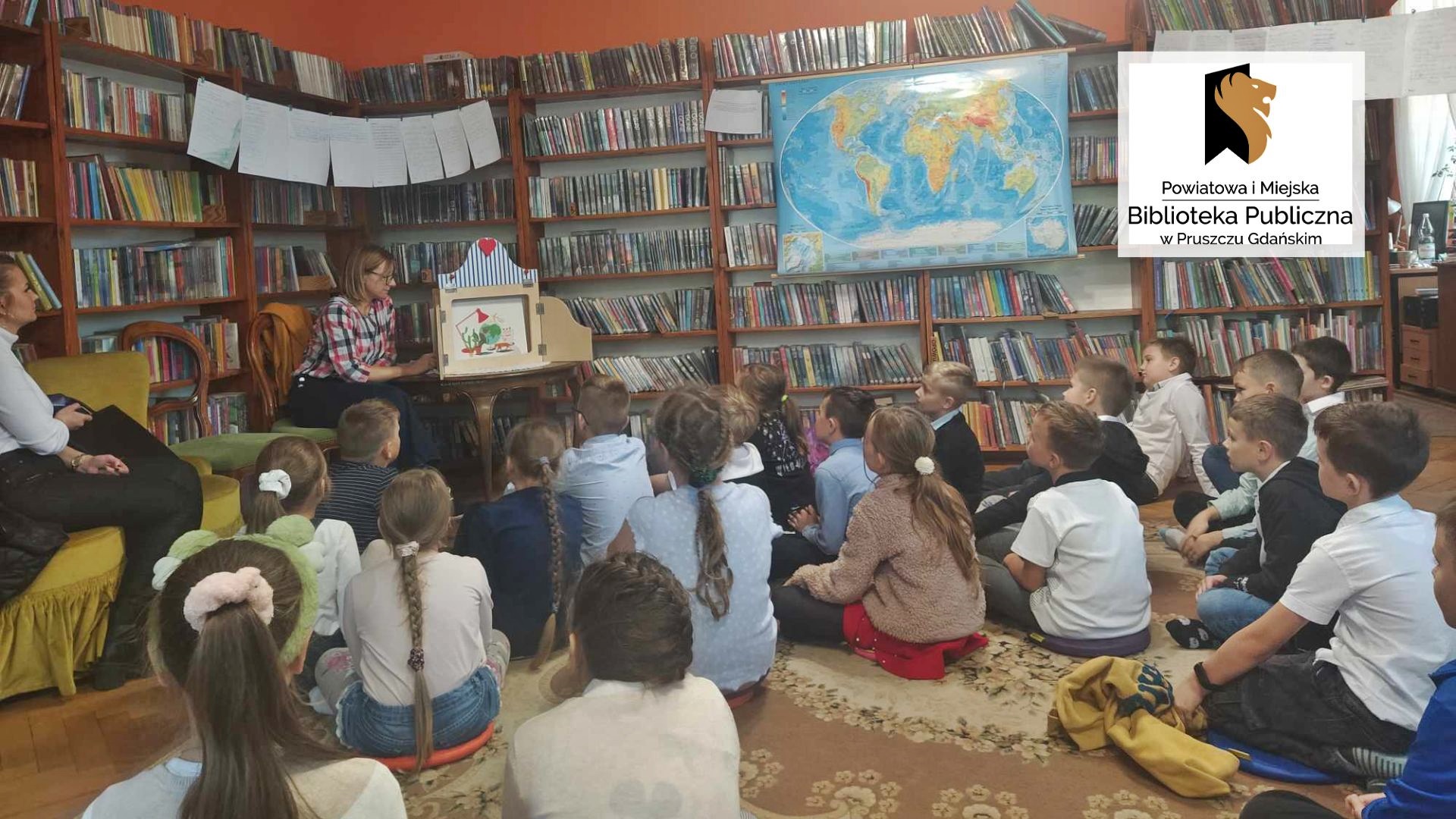 Dzieci siedzą na podłodze i patrzą w kierunku teatrzyku kamishibai, który stoi na stoliku. W teatrzyku znajduje się kolorowa ilustracja. Po lewej siedzi bibliotekarka, a obok niej nauczycielka. Po prawej wisi mapa świata. W tle regały z książkami.
