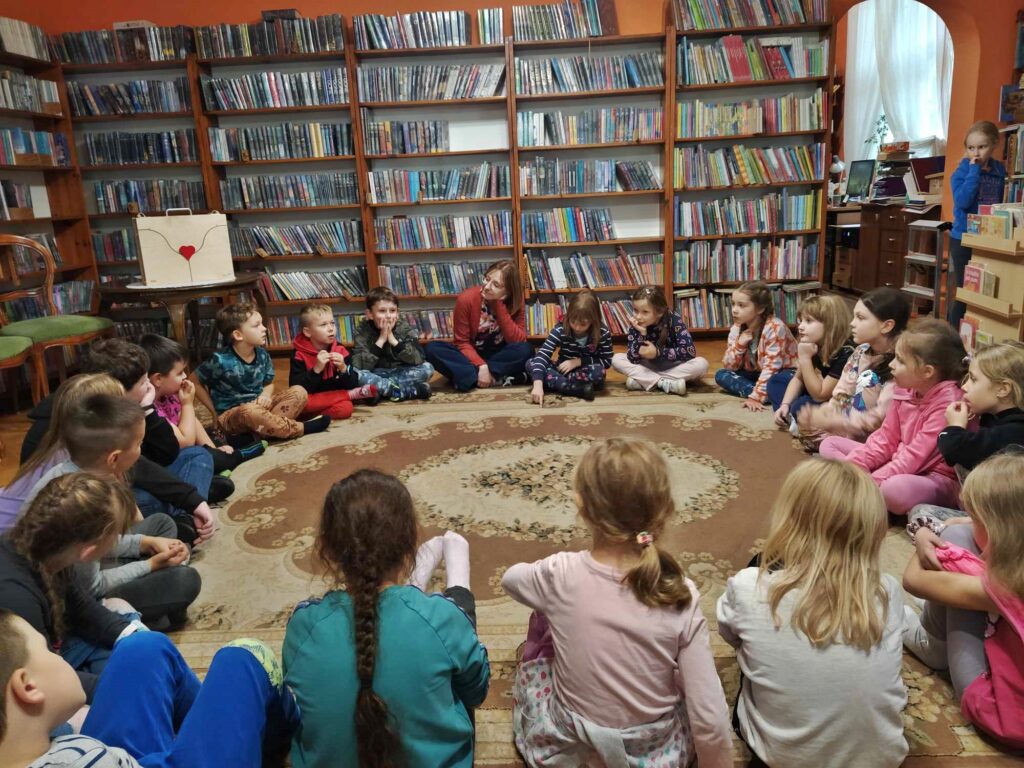 Dzieci siedzą na podłodze i patrzą w stronę teatrzyku kamishibai, który stoi na stoliku. Po lewej siedzi bibliotekarka i czyta. W tle regały z książkami.